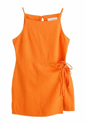 Frau mit dunklen Haaren, die einen ärmellosen orangefarbenen Overall trägt, steht draußen neben einer weißen Wand und grünen Pflanzen, seitlich blickend.