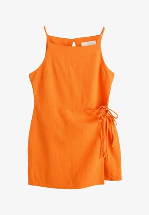 Žena s tmavými vlasmi, ktorá nosí oranžový overal bez rukávov, stojí vonku blízko bielej steny a zelených rastlín, otočená bokom.