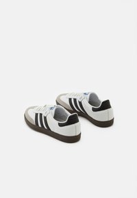 Zapatillas blancas con detalles en ante gris, rayas negras y una suela marrón texturizada. Los cordones son destacados, con un detalle del logo azul en la lengüeta.