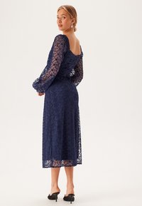 Mörkblå spetsklänning med långa ärmar, blommönster, rynkad bak och midi-längd. Bärs med svarta klackar; genomskinliga detaljer på ärmarna.