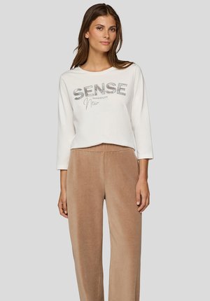 Frau trägt weißes Langarmshirt mit dem Schriftzug "SENSE" und beigefarbene Cordhosen, steht vor einem einfarbigen grauen Hintergrund.
