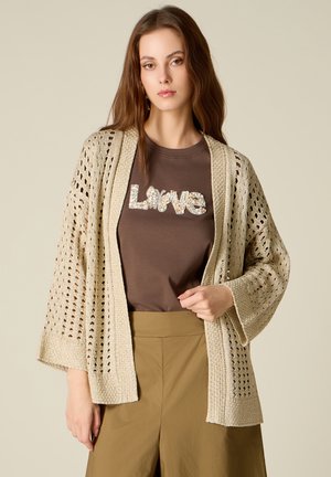Giovane donna con un cardigan beige a maglia aperta sopra una maglietta marrone decorata con la scritta "Love" e pantaloni kaki, in piedi su uno sfondo semplice.