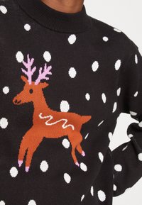 Pull noir avec des pois blancs, un motif de renne orange avec des bois roses et un accent ondulé blanc sur le côté.