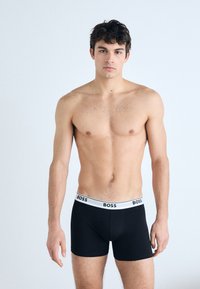 Schwarze Boxer-Shorts mit einem weißen Bund, der das "BOSS"-Logo trägt. Das Modell trägt kein Oberteil und steht vor einem hellen Hintergrund.