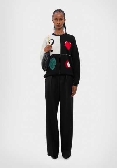 Maglione nero a maglia con motivi colorati: cuore, foglia, frutto e punto interrogativo. Indossato con pantaloni neri larghi e portando una borsa nera.