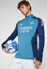 Blue and navy athletic jersey with a half-zip collar, featuring "Emirates Fly Better" logo and a textured side panel. Holding a patterned soccer ball.