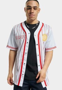 Tricou de baseball alb cu dungi roșii, mâneci scurte, nasturi de tip snap și un grafic cu soare. Purta peste un tricou negru, cu text brodat.