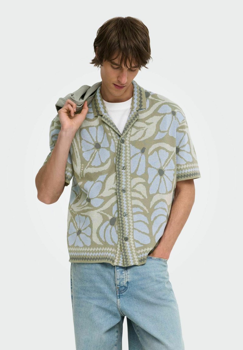 Jeune homme portant une chemise à manches courtes boutonnée avec un motif floral bleu, un jean clair et tenant une veste sur son épaule.