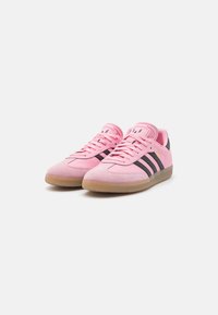 Pink suede sneakers with black stripes and logo, featuring a textured rubber sole and lacing system. Classic design with rounded toe.
