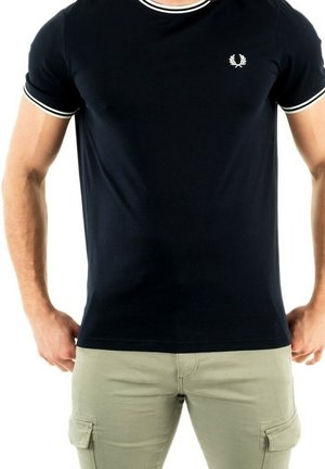 Homme portant un t-shirt noir ajusté avec des bordures blanches aux manches et au col, associé à un pantalon cargo beige clair, debout les mains le long du corps.