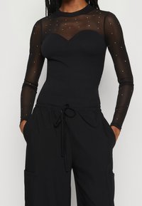 Svart långärmad topp med ett genomskinligt, prickigt överdrag och en hjärtformad halsringning. Matchas med svarta löst sittande byxor med fickor.