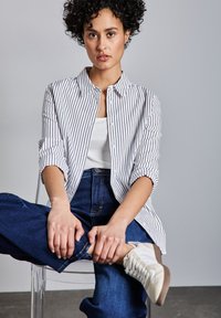 Camicia leggera a righe verticali bianca e nera, abbinata a jeans denim blu scuro e sneakers color crema. Modello seduto su una sedia.