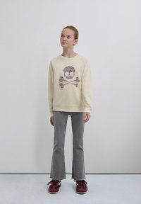 Sweat-shirt de couleur crème avec un design coloré de crâne et os croisés, assorti à un jean évasé gris et des baskets bordeaux.