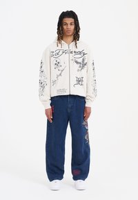 Sudadera beige con cremallera y estampados gráficos; vaqueros anchos azules con detalles bordados; zapatillas blancas; exhibidos sobre un fondo liso.