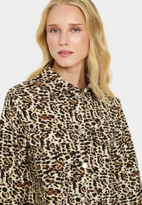 Bežinė ir juoda leopardų rašto marškinėlių striukė su mygtukų užsegimu, dviem krūtinės kišenėmis ir apykakle; pagaminta iš tekstūruoto audinio.