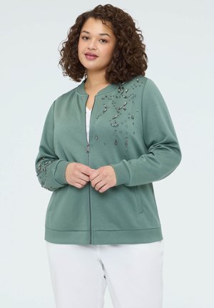 Kvinde med krøllet hår iført en grøn lynlåstrøje dekoreret med perler på skulderen og ærmet, kombineret med hvide bukser.