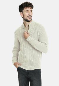 Suéter de punto beige claro con cuello acanalado, que presenta un diseño de trenzas y mangas largas. Lleva puesto sobre una camisa color canela y combinado con jeans oscuros.