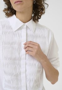 Chemise blanche à col avec des plis verticaux à l'avant. Tissu lisse, fermeture à boutons et manches retroussées. Cheveux bouclés partiellement visibles.