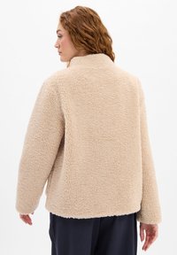 Frau mit welligem roten Haar, die eine beigefarbene, flauschige Fleecejacke trägt, von hinten vor einem schlichten weißen Hintergrund betrachtet.