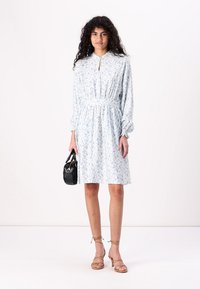 Vestido de estampado floral en azul claro y blanco, con cuello, cintura elástica, mangas abullonadas largas y tejido suave. Sostenido con un pequeño bolso negro.
