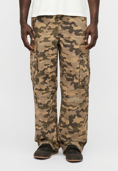 Pantalon cargo camouflage en tons beige et vert, en coton. Il comporte des poches latérales, une coupe décontractée et une zone usée près du genou.