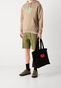 Felpa con cappuccio tan e logo verde, pantaloni corti verde oliva, borsa a tracolla nera con logo rosso, sneakers verdi e calzini neri con dettagli del marchio.