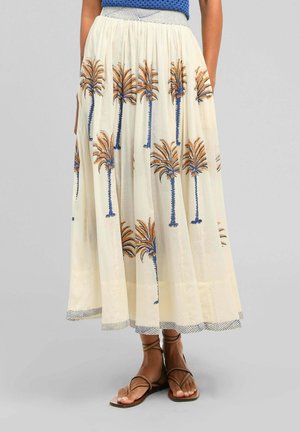Jupe crème claire avec des imprimés de palmiers bleus et orange, portée avec des sandales marron à brides sur une personne les mains dans les poches.