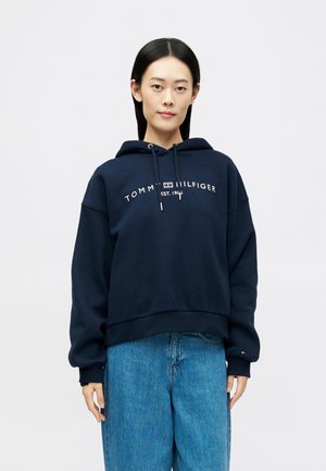 Jeune personne avec des cheveux noirs raides portant un sweat à capuche Tommy Hilfiger bleu marine et un jean bleu sur un fond uni.