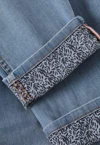 Țesătura de denim cu o nuanță albastră, având o manșetă imprimată în marin închis cu un model de frunze, accentuată de cusături portocalii pe acele cusături.