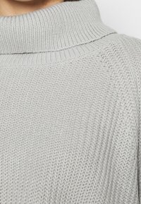 Ljust grått stickat tröja med hög, ribbad krage. Texturen är mjuk, med en lös väv och subtila mönster genom hela plagget.