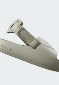 Adidas sandal v svetlo sivi barvi z nastavljivim trakom, teksturirano površino in nizkim profilom. Ima logotip vtisnjen na strani.