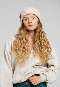 Kremowa czapka z ribbowanym splotem, z skórzanym akcentem, w połączeniu z beżowym polarowym zamkiem błyskawicznym i niebieskimi dżinsami, charakteryzująca się miękkimi, kręconymi włosami.