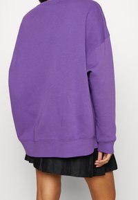 Purple oversized sweatshirt with ribbed cuffs and hem, made of soft fabric, paired with a black pleated skirt, viewed from the back.