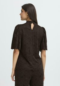 Blusa in pizzo floreale marrone con maniche a sbuffo corte, scollatura alta e apertura a chiave nella parte superiore della schiena. Motivo strutturato lungo tutta la superficie.