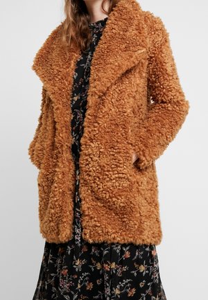 Mujer con un abrigo de piel sintética rizada de longitud media en color naranja-marrón sobre un vestido negro con estampado floral, con una mano en el bolsillo del abrigo.