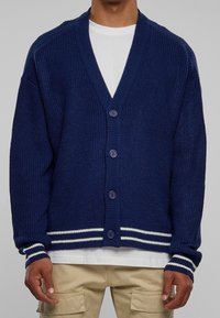 Marineblauw vest met een getextureerd breisel, v-hals, sluiting met zes knopen en witte gestreepte accenten op de manchetten en zoom. Gedragen over een wit shirt.