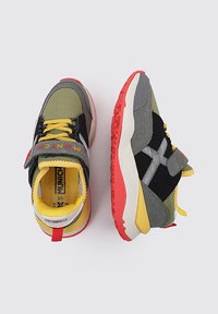 Las zapatillas deportivas presentan un upper de malla y sintético en verde, negro y gris, con un interior amarillo, suela roja y correa de velcro.