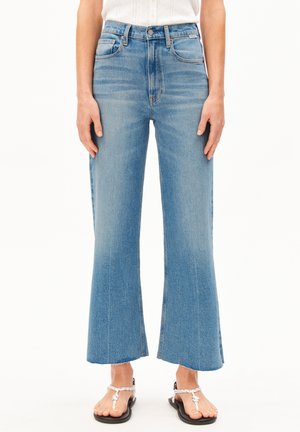 Vue de face d'une personne portant un jean évasé court taille haute bleu clair avec des ourlets bruts et des sandales à brides blanches tressées.