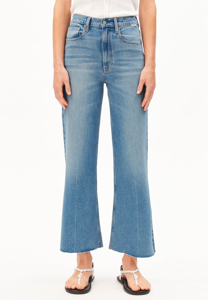 Vorderansicht einer Person, die hellblaue hoch taillierte, gekürzte Schlagjeans mit rohen Säumen und weißen Sandalen mit geflochtenen Riemen trägt.