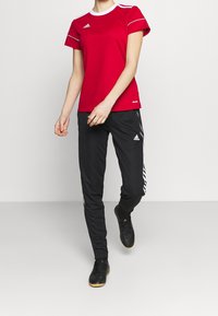 Red athletic t-shirt with white accents and logo, paired with black pants featuring side stripes and mesh details, worn with black sports shoes.