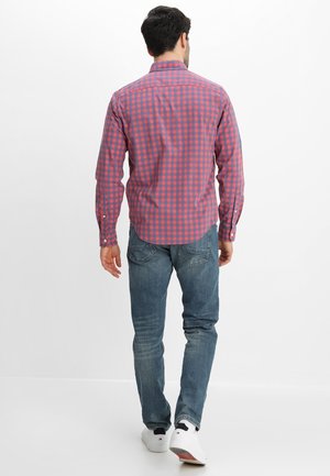Mann, der ein rot-blau kariertes Langarmhemd, blaue Jeans und weiße Turnschuhe trägt, läuft auf weißem Hintergrund weg.