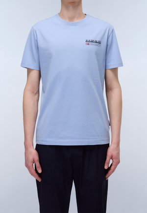 Mand iført en lyseblå Napapijri t-shirt med korte ærmer, lille logo på brystet og sorte bukser foran en ensfarvet baggrund.