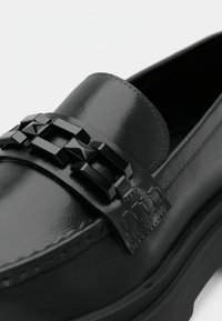 Mocassim preto em pele com acabamento brilhante, com um acessório de corrente geométrica preta na pala e detalhe de costura texturizada nas laterais.