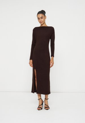 Femme avec les cheveux en chignon, portant une robe marron foncé à manches longues avec une fente à l'avant et des sandales à talons hauts noires à brides, debout devant un fond blanc.