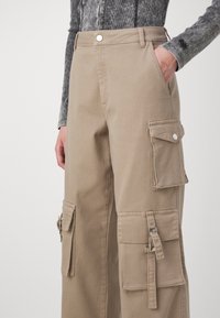 Personne portant un pantalon cargo beige avec plusieurs poches, comprenant des rabats et des détails de boucle, associé à un haut gris à manches longues.