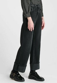 Persoon die een paar brede zwarte jeans met manchet draagt, zwarte gestructureerde sokken, zwarte leren instappers met gesp en een zwart-wit geblokt overhemd.