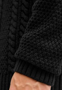 Zwarte gebreide trui in close-up, met textuurpatronen, geribde manchetten en een chunky ontwerp. De stof lijkt zacht en dik.