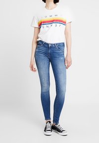 Vit t-shirt med färgglada horisontella ränder och texten "TOMMY JEANS"; blå skinny jeans; svarta sneakers med vita snören.