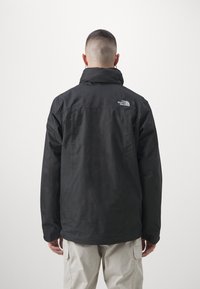 Personne aux cheveux courts portant une veste noire The North Face et un pantalon clair, vue de dos sur un fond clair uni.