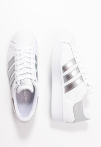 Bílé kožené tenisky se stříbrnými akcenty, s texturovanou špičkou, třemi kovovými pruhy a plochou gumovou podrážkou.